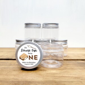 May include: A set of clear glass jars with silver lids. The jars are decorated with a round sticker that says "It's S'more Fun being One" with a graphic of a s'more. The sticker also says "Milo's 1st Birthday August 6, 2025".
