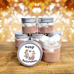 May include: A set of six small glass jars with silver lids filled with hot chocolate mix and marshmallows. The jars are stacked in a pyramid shape. The jar on top has a round label that says "Happy Halloween" with a ghost and flowers.