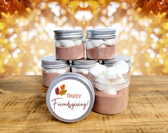 May include: A stack of six clear glass jars with silver lids filled with hot chocolate mix and marshmallows. The jars are labeled with a white sticker that says "Happy Friendsgiving!" with a fall leaf design.