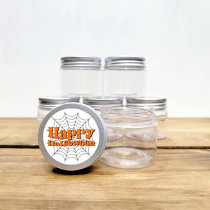 May include: A set of six clear glass jars with silver lids. One jar has a round sticker with a black spiderweb and the text "Happy Halloween" in orange.