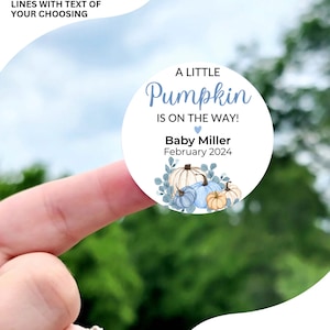 May include: A round sticker with a blue and white pumpkin design. The text reads "A Little Pumpkin Is On The Way!" with a heart symbol. Below the text is "Baby Miller February 2024". The sticker is available in sizes 1 inch, 1.5 inches, 1.75 inches, 2 inches, 2.25 inches, and 3.33 inches.