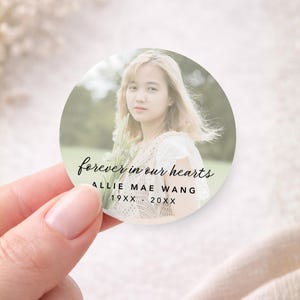 May include: A round sticker featuring a photograph of a woman holding flowers. The text "forever in our hearts" is written in cursive, along with the name "Allie Mae Wang" and dates. The sticker is held by a hand.