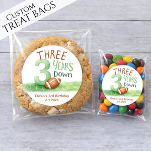 May include: Two clear treat bags, one with a large cookie and the other with colorful candies. Both bags have a circular sticker with the text "THREE YEARS DOWN" and a football graphic. The text "CUSTOM TREAT BAGS" is at the top.