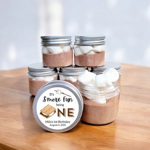 May include: Silver jars filled with chocolate powder, marshmallows, and graham crackers. The jars have labels that say "It's S'more Fun being ONE" and "Milo's 1st Birthday August 6, 2025".