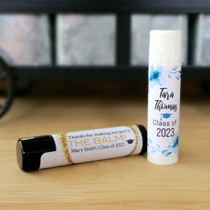 May include: Two graduation party favors, lip balm tubes with personalized labels. One label features a blue floral design with the text "Tara Thomas Class of 2023". The other label has a gold glitter background with the text "Thanks for making my party THE BALM! Mary Smith | Class of 2023".