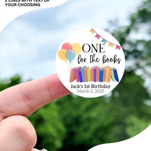 May include: A round sticker with a white background and colorful illustrations. The sticker says "ONE for the books" with a stack of colorful books and balloons. The text below the illustration says "Jack's 1st Birthday March 2, 2025". The sticker is available in sizes 1 inch, 1.5 inches, 1.75 inches, 2 inches, 2.25 inches, and 3.33 inches. The sticker pictured is 2 inches.