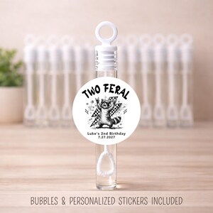 May include: Clear bubble wand with a white cap and a round sticker. The sticker features a black and white illustration of a raccoon with the text "TWO FERAL" and "Luke's 2nd Birthday 7.27.2027". The background shows more bubble wands.