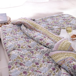 Op de afbeelding: Een bloemmotief quilt in wit, paars en blauw, met een contrasterende rand. De quilt ligt op een bed, met een boek, een glas thee en koekjes zichtbaar. Het ontwerp van de quilt toont bloemen en vogels. Afmetingen in cm.