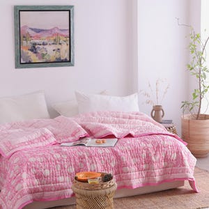 May include: A pink and white patterned quilt adorns a bed, paired with white pillows. A small woven side table holds a tray with fruit and a pinecone. A framed landscape painting hangs on the wall, adding an artistic touch.