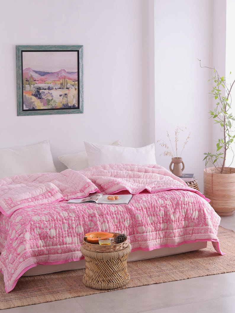 May include: A pink and white patterned quilt adorns a bed, paired with white pillows. A small woven side table holds a tray with fruit and a pinecone. A framed landscape painting hangs on the wall, adding an artistic touch.
