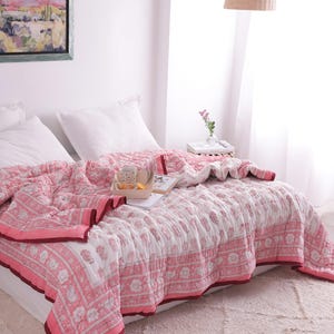 May include: A pink and white floral patterned quilt covers a bed, with a matching border and a dark red trim. White pillows and a tray with a cup and a snack sit on the bed. A framed landscape painting hangs on the wall.