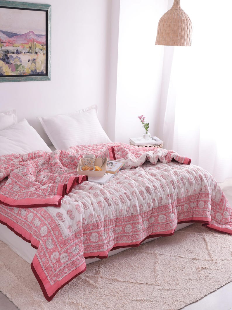 May include: A pink and white floral patterned quilt covers a bed, with a matching border and a dark red trim. White pillows and a tray with a cup and a snack sit on the bed. A framed landscape painting hangs on the wall.