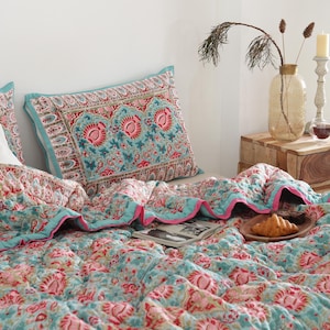 Może przedstawiać: Łóżko z kołdrą we wzory kwiatowe i pasującymi poszewkami w odcieniach turkusu, różu i bieli. Drewniana szafka nocna zawiera wazon z suszonymi roślinami, świecę i szklankę płynu. Rogalik spoczywa na drewnianym talerzu.