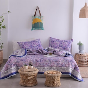 May include: A bedroom scene featuring a floral patterned quilt in shades of purple and blue, with matching pillowcases. Two small woven stools and a tote bag with a Nashville skyline are also visible. A potted plant and a woven pendant light complete the decor.