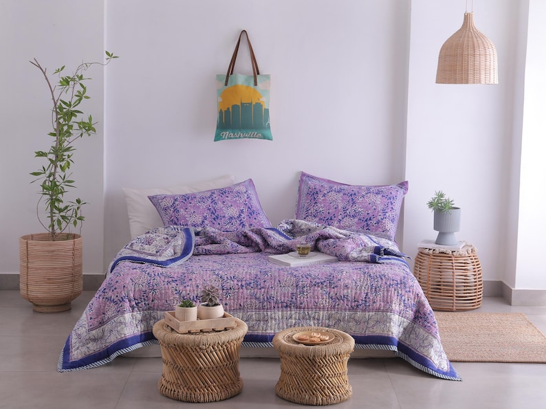 May include: A bedroom scene featuring a floral patterned quilt in shades of purple and blue, with matching pillowcases. Two small woven stools and a tote bag with a Nashville skyline are also visible. A potted plant and a woven pendant light complete the decor.