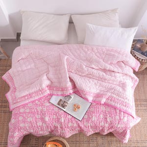 May include: A bed is shown from above, featuring a pink and white patterned quilt and two white pillows. The quilt has a floral design on the lower section and a striped pattern on the upper. A woven rug and a small side table with a plant are also visible.