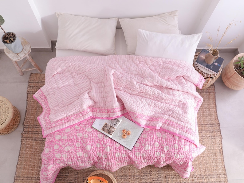 May include: A bed is shown from above, featuring a pink and white patterned quilt and two white pillows. The quilt has a floral design on the lower section and a striped pattern on the upper. A woven rug and a small side table with a plant are also visible.