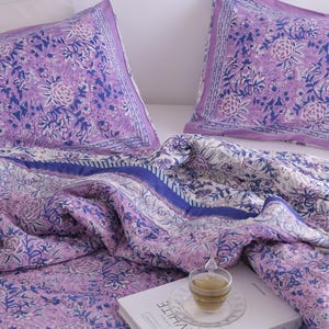 May include: A bedding set featuring a floral pattern in shades of lavender, white, and blue. The set includes two pillowcases and a quilt, with a book and a cup of tea on top. The word "WHITE" is visible on the book.