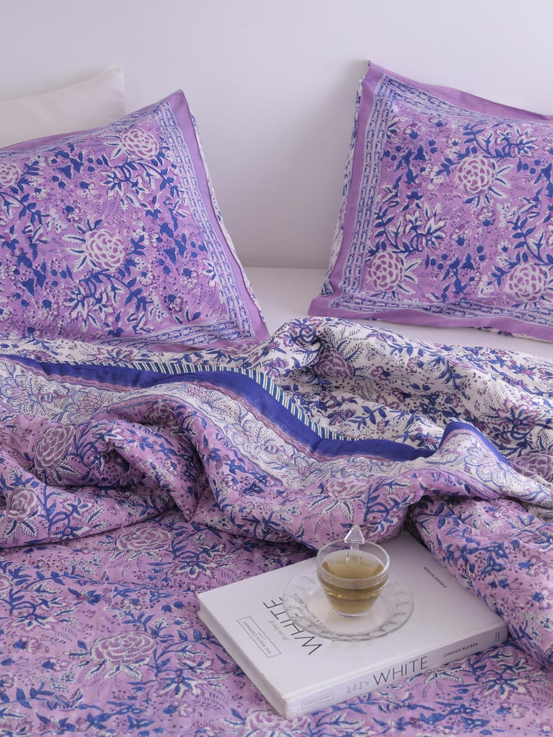 May include: A bedding set featuring a floral pattern in shades of lavender, white, and blue. The set includes two pillowcases and a quilt, with a book and a cup of tea on top. The word "WHITE" is visible on the book.