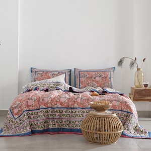 May include: A floral patterned quilt set in shades of coral, blue, and green, with matching pillowcases. The bedding is displayed on a bed with a white wall backdrop. A small wooden side table and a woven side table are also in the image.
