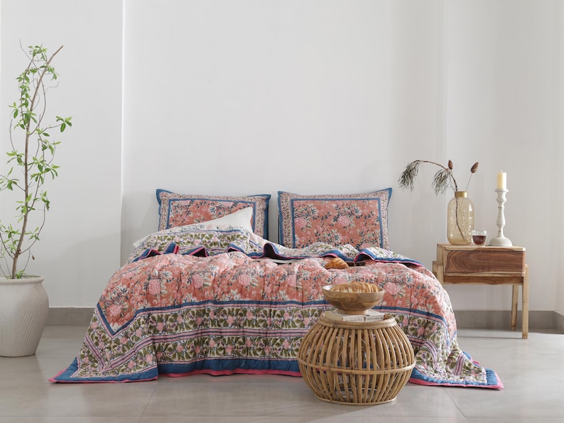 May include: A floral patterned quilt set in shades of coral, blue, and green, with matching pillowcases. The bedding is displayed on a bed with a white wall backdrop. A small wooden side table and a woven side table are also in the image.
