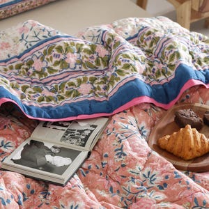 May include: A floral patterned quilt in shades of pink, blue, and green, with a blue border and pink trim. An open book and a wooden plate with a croissant and two brownies rest on the quilt.