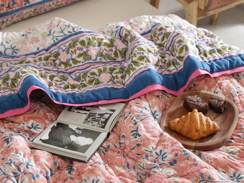 May include: A floral patterned quilt in shades of pink, blue, and green, with a blue border and pink trim. An open book and a wooden plate with a croissant and two brownies rest on the quilt.