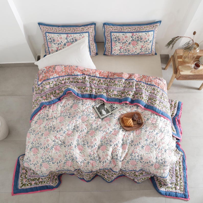 May include: A floral patterned quilt and two matching pillowcases in pink, blue, and green hues. The quilt features a repeating flower design on a white background, with a blue border and pink trim. A white pillow is on the bed.