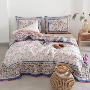 May include: A floral patterned quilt and two matching pillowcases in pink, blue, and white. The quilt features a repeating floral design with a blue border and pink trim. The pillowcases have a similar floral pattern with a blue border. The bedding is on a bed with a white pillow.