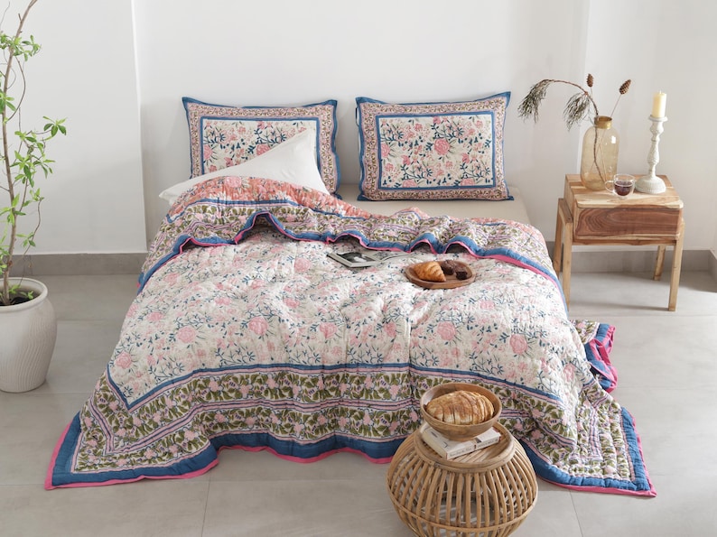 May include: A floral patterned quilt and two matching pillowcases in pink, blue, and white. The quilt features a repeating floral design with a blue border and pink trim. The pillowcases have a similar floral pattern with a blue border. The bedding is on a bed with a white pillow.