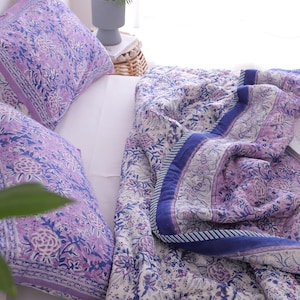 May include: A bed set featuring a floral patterned quilt and two matching pillowcases. The bedding has a white background with a purple and blue floral design. The quilt has a blue border. A cup of tea and a book are on the bed.