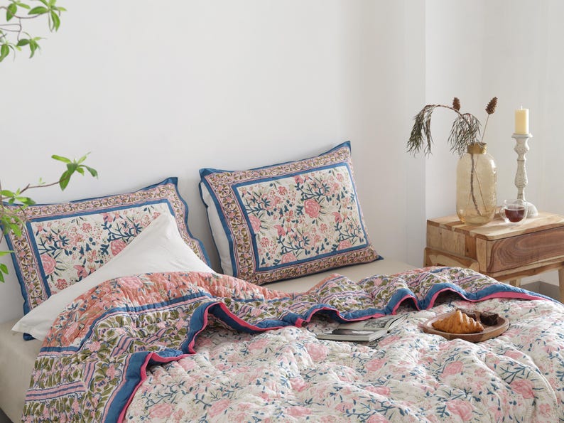 May include: A bed with a floral patterned quilt and two matching pillowcases. The quilt features pink and green floral designs on a white background, with blue and pink borders. A white pillow is placed between the pillowcases.