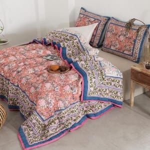 May include: A bed is covered with a floral patterned quilt and matching pillow shams. The quilt is primarily peach with pink and green floral designs, bordered by blue and pink trim. Two matching pillow shams are on the bed. A small wooden side table holds a candle and a vase.