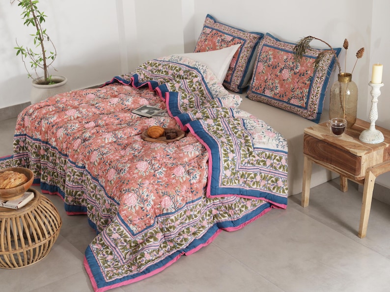 May include: A bed is covered with a floral patterned quilt and matching pillow shams. The quilt is primarily peach with pink and green floral designs, bordered by blue and pink trim. Two matching pillow shams are on the bed. A small wooden side table holds a candle and a vase.