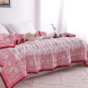 May include: A bed is covered with a pink and white floral patterned quilt. The quilt has a dark pink border and is draped over white pillows and a mattress. A wooden tray with items sits on the quilt. A vase with flowers is on the bed.