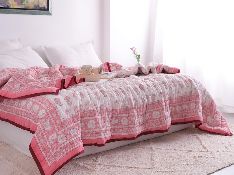 May include: A bed is covered with a pink and white floral patterned quilt. The quilt has a dark pink border and is draped over white pillows and a mattress. A wooden tray with items sits on the quilt. A vase with flowers is on the bed.
