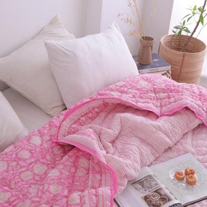 May include: A bed is made with white pillows and a pink patterned quilt. The quilt has a floral design on the top layer and a striped pattern on the underside. A book and a plate of pastries are on the bed.