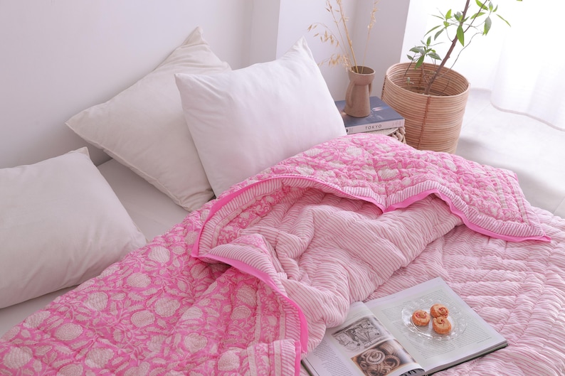 May include: A bed is made with white pillows and a pink patterned quilt. The quilt has a floral design on the top layer and a striped pattern on the underside. A book and a plate of pastries are on the bed.
