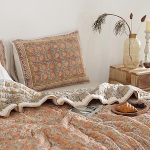 May include: A bed is made with a floral patterned quilt and matching pillowcases in shades of orange, green, and white. A wooden nightstand holds a vase with dried plants, a candle, and a small glass. A wooden tray with a croissant and a pastry sits on the quilt.