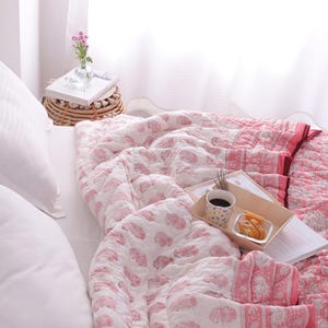 May include: A bed with white pillows and a pink and white patterned quilt. A wooden tray holds a cup of coffee, snacks, and a book. A small side table with a vase of flowers sits nearby.