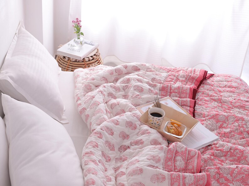 May include: A bed with white pillows and a pink and white patterned quilt. A wooden tray holds a cup of coffee, snacks, and a book. A small side table with a vase of flowers sits nearby.