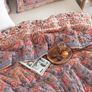 May include: A bed with a patterned quilt in shades of gray, red, and white. The quilt features floral and paisley designs. Pillows with matching patterns are visible. A wooden tray with snacks and an open book are on the quilt.