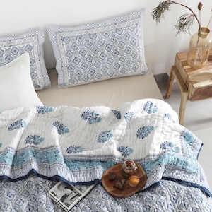 May include: A bed featuring a white and blue floral quilt and pillowcases. The quilt has a blue border and floral patterns. A wooden side table holds a vase, candle holder, and a small tray with treats. A book and a wooden tray with pastries are on the bed.