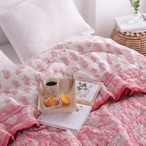 May include: A bed is made with a white pillow and a pink and white paisley patterned quilt. A wooden tray holds a cup of coffee, a pastry, and an orange slice. A small woven side table holds a book and a vase of flowers.