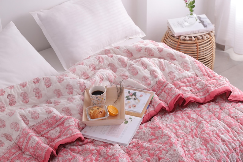 May include: A bed is made with a white pillow and a pink and white paisley patterned quilt. A wooden tray holds a cup of coffee, a pastry, and an orange slice. A small woven side table holds a book and a vase of flowers.