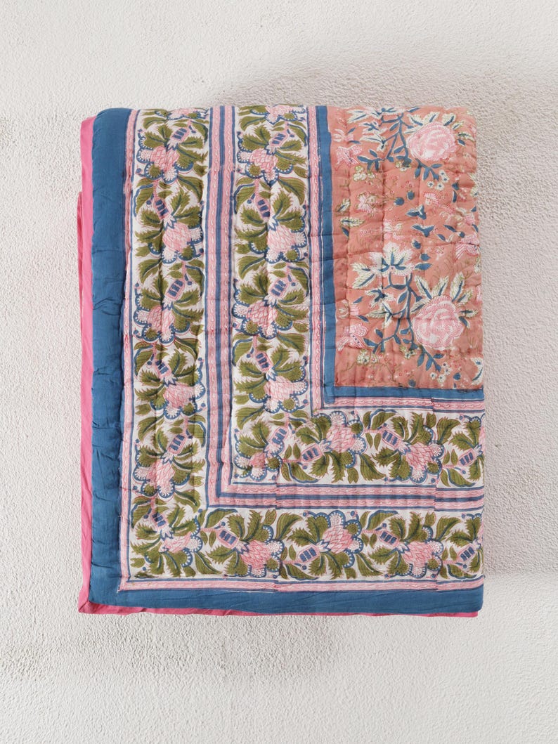 May include: A folded, quilted blanket with a floral pattern. The blanket features a coral-colored center with pink roses and blue leaves. The border has a repeating floral design in pink, green, and blue. The edges are trimmed with blue and pink fabric.