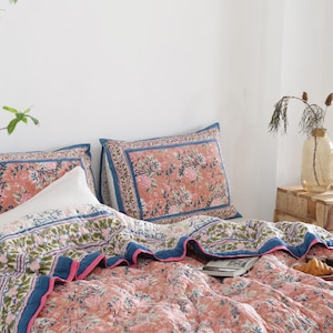 May include: A bed is made with a floral patterned quilt and matching pillowcases. The quilt and pillowcases have a coral background with pink and green floral designs, and blue borders. A white pillow is also on the bed.