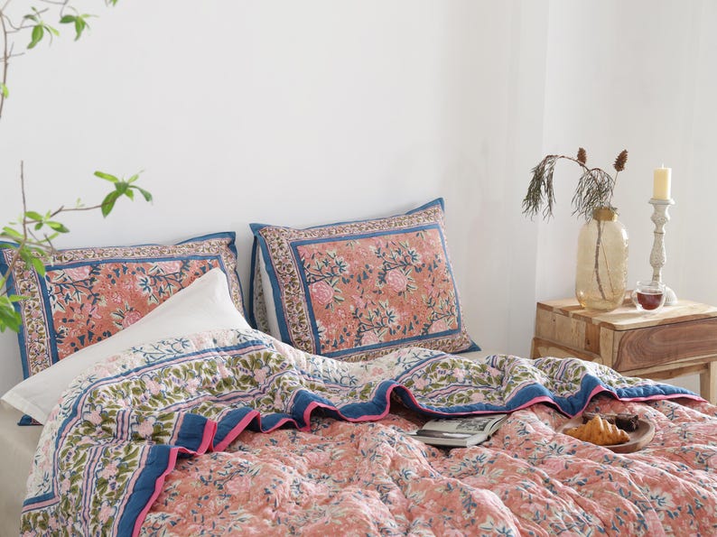 May include: A bed is made with a floral patterned quilt and matching pillowcases. The quilt and pillowcases have a coral background with pink and green floral designs, and blue borders. A white pillow is also on the bed.