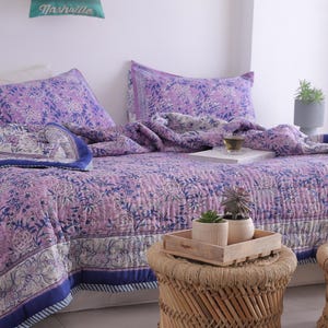 May include: A bed is made with a purple floral patterned quilt and matching pillowcases. The quilt has a blue border. A small wooden tray with plants sits on a woven stool. A pillow with the word "Nashville" is on the wall.