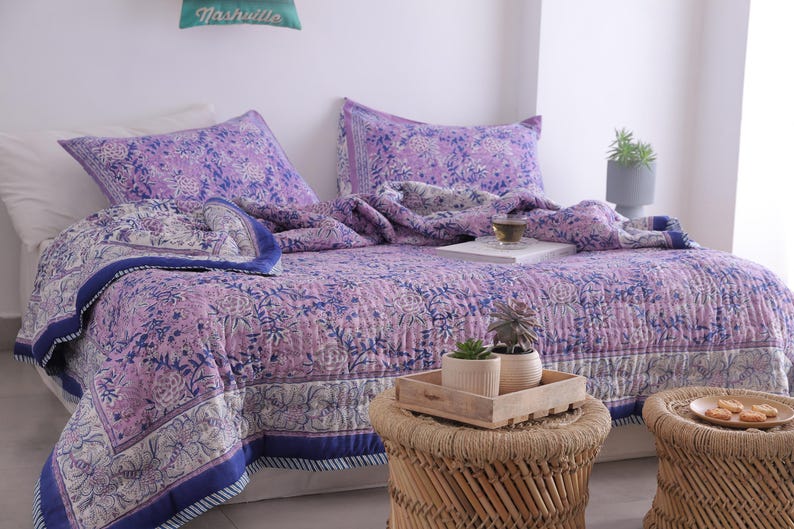 May include: A bed is made with a purple floral patterned quilt and matching pillowcases. The quilt has a blue border. A small wooden tray with plants sits on a woven stool. A pillow with the word "Nashville" is on the wall.